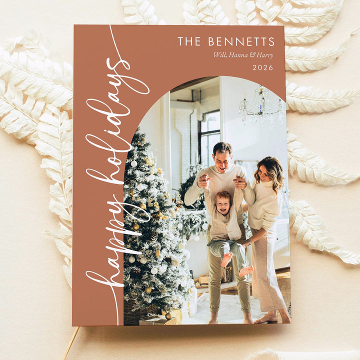 Close-up of a terracotta Happy Holidays family photo card with white script, personalized names, and an arched photo layout.
