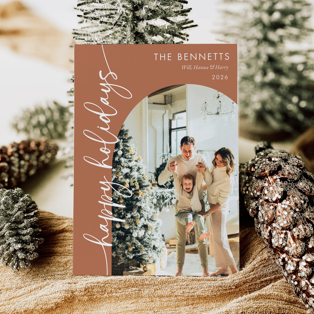 Terracotta holiday photo card with white Happy Holidays script and an arched family portrait, displayed among Christmas trees and pinecones.