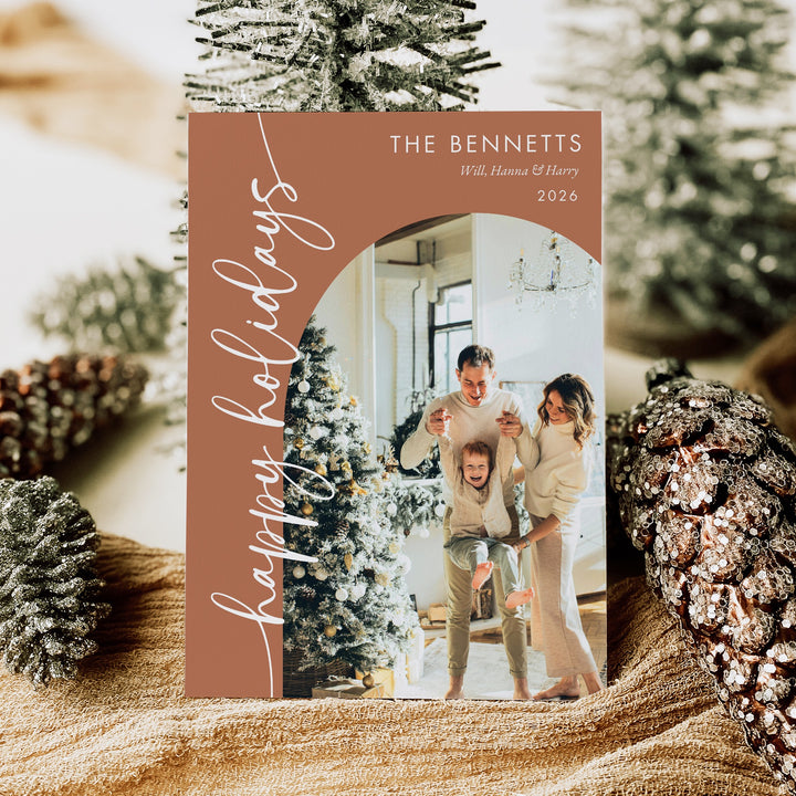Terracotta holiday photo card with white Happy Holidays script and an arched family portrait, displayed among Christmas trees and pinecones.